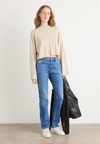Beige avkortet langermet skjorte, blå rette jeans, hvite sneakers og en svart skinnjakke holdt i høyre hånd.