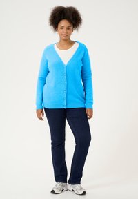Hellblauer, flauschiger Cardigan mit V-Ausschnitt, durchgehender Knopfleiste und langen Ärmeln, kombiniert mit dunkelblauen Jeans und grauen Sneakers.