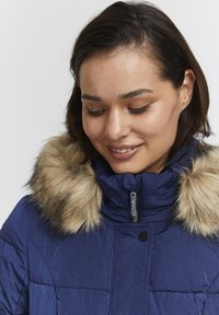 Mörkblå puffjacka med fuskpälskrage, som har en hög dragkedja i halsen, quiltad textur och tryckknappsstängning.