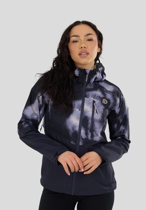 Mujer con pelo largo y oscuro, vistiendo una chaqueta negra y morada con cremallera y estampado de nubes, con capucha, de pie frente a un fondo liso.