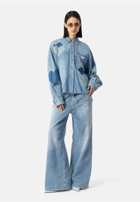 Žena v prevelkej svetlomodrej džínovej košeli s nášivkami srdca a širokými džínami, stojí s jednou rukou v vrecku, na neutrálnom pozadí.