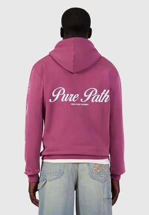 Man wearing a purple hoodie with "Pure Path" and "Own Your Journey" text on the back, paired with light blue denim jeans.