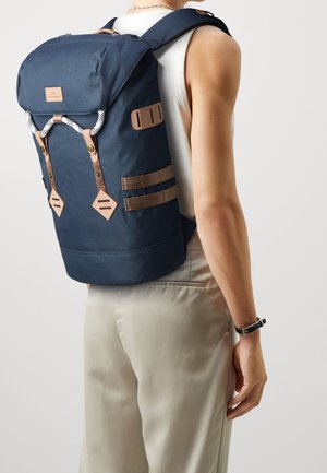 Sac à dos bleu marine avec rabat supérieur, accents en cuir et bretelles réglables. Comprend des connecteurs en forme de diamant et une surface texturée.