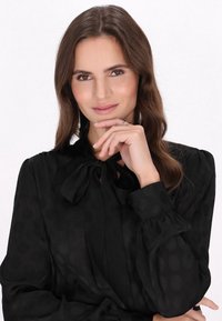 Sort bluse med sløjfe ved halsen, lange pufærmer og tekstureret prikmønster. Model poserer med hånden på hagen.
