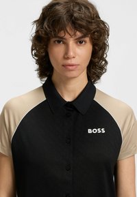Svart och beige polo skjorta med krage, korta ärmar och textilstruktur. Varumärket "BOSS" visas i vitt.
