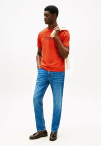 Camiseta de algodón roja con logo, jeans de mezclilla azules y mocasines marrones. El modelo sostiene una toalla blanca colgada sobre el hombro. Atuendo simple y casual.