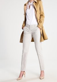 Cappotto trench beige su una camicia bianca con bottoni, abbinato a pantaloni slim-fit grigio chiaro texturizzati e tacchi alti color nude, creando un look a strati.