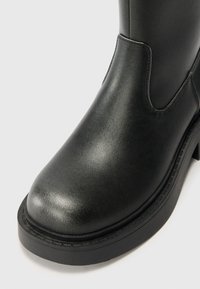 Gros plan d'une botte en cuir noire avec un bout arrondi et une semelle épaisse sur un fond clair uni.