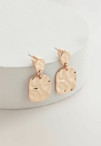 Roségoldene Ohrringe mit einem strukturierten, unregelmäßigen rechteckigen Anhänger und einer runden Steckerbefestigung, präsentiert auf einem kreisrunden weißen Sockel.