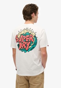 Hvid bomulds t-shirt med en farverig grafisk design af ordene "SUPER DRY" omkranset af bølgemønstre på ryggen.