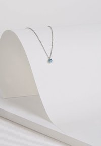 Zilveren ketting met een ronde blauwe edelsteen hanger, ingesteld in een eenvoudige zilveren sluiting. Tentoongesteld op een gebogen witte ondergrond.