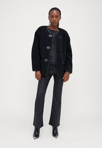 Čierna flísová bunda s voľným strihom, okrúhlym výstrihom a zapínaním na šnúrky, kombinovaná s čiernymi nohavicami z umelej kože a čiernymi špicatými členkovými topánkami.