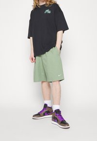 Svart oversized t-shirt med grön logotyp, gröna shorts och bruna högkottade sneakers med lila snören. Tillbehör inkluderar vita strumpor.