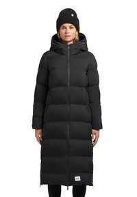 Cappotto imbottito nero con un design lungo, motivo a righe orizzontali, zip frontale e due tasche laterali. Indossato con un berretto di maglia nero.