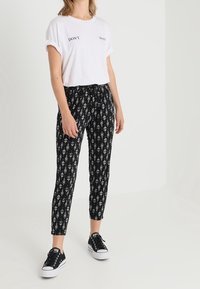 Witte T-shirt met "DON'T WANT" in het zwart gedrukt, gecombineerd met zwarte geblokte broek met witte geometrische ontwerpen, gedragen met zwarte sneakers.