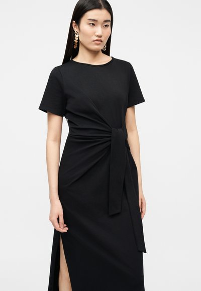 Γυναίκα που φοράει μαύρο φόρεμα με κοντά μανίκια, κόμπο μπροστά και σκίσιμο στο πλάι, διακοσμημένο με μεγάλα χρυσά σκουλαρίκια σε σχήμα δίσκου.