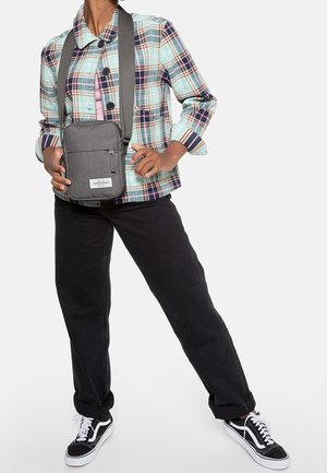 Person trägt eine kariertes Jacke, schwarze Hose, schwarz-weiße Turnschuhe und eine graue Umhängetasche mit Eastpak-Logo.