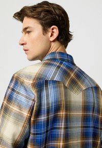 Camisa de cuadros con colores azul, marrón y crema. Confeccionada en una tela ligera con cuello pointed y un diseño de hombros en contraste.