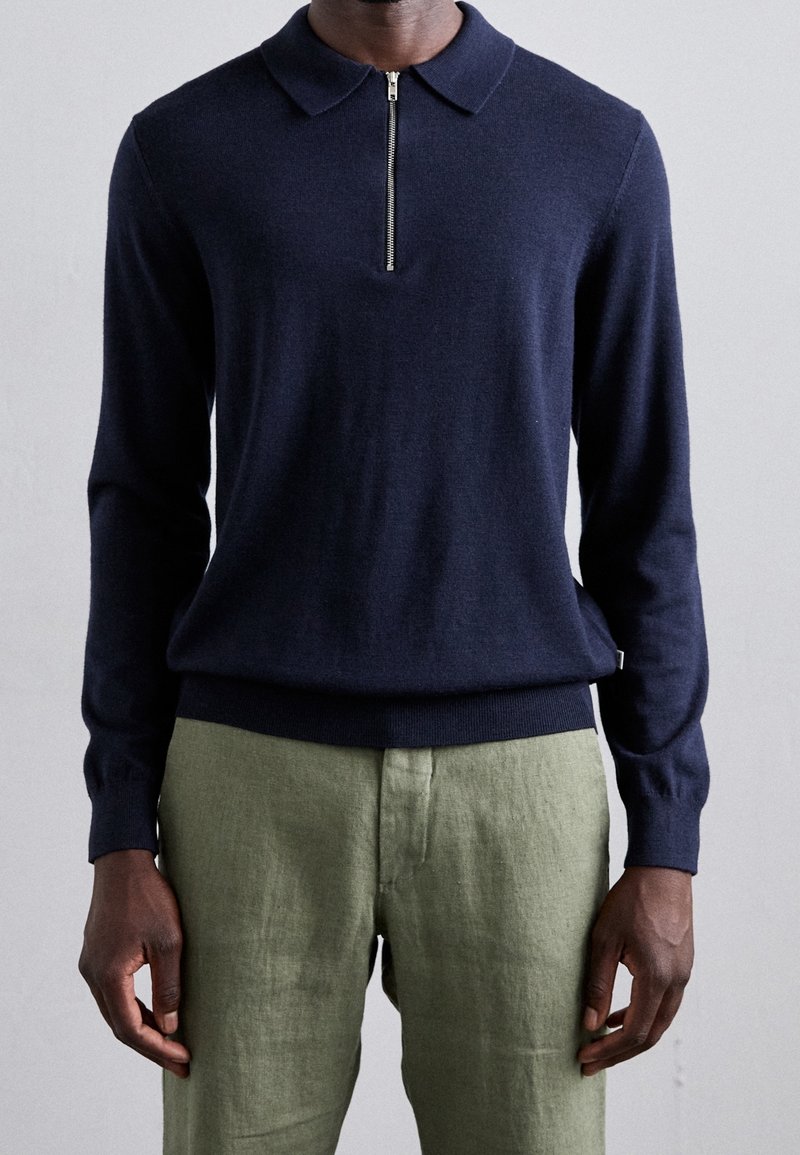 Mörkblå stickad tröja med dragkedja i kragen, ribbad mudd och midja, matchad med ljusgröna linbyxor. Texturerad tyg med en slät yta.