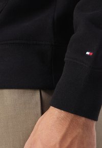 Sort sweatshirt med ribbet manchetter; har et lille logo med rød, hvid og blå striber. Hånd hviler på lysebrune bukser.