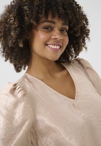 Bloedroze, gestructureerde blouse met een v-hals en pofmouwen. Het materiaal heeft een lichte glans, die zachte plooien en opzettelijke kreukels toont.
