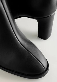 Schwarze, knöchelhohe Stiefel mit Absatz aus strukturiertem Leder, mit spitz zulaufender Zehenpartie, glatten, geschwungenen Kanten und robustem Blockabsatz.