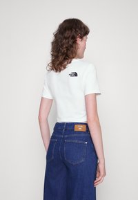 Camiseta blanca de manga corta con un logo negro en la parte trasera, combinada con unos vaqueros de mezclilla oscuros de tiro alto que presentan una etiqueta marrón.
