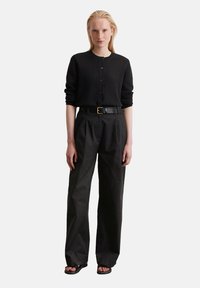 Schwarze Strickjacke mit Knöpfen, kombiniert mit locker sitzenden schwarzen Hosen und einem schwarzen Gürtel mit goldener Schnalle, getragen mit flachen Sandalen.