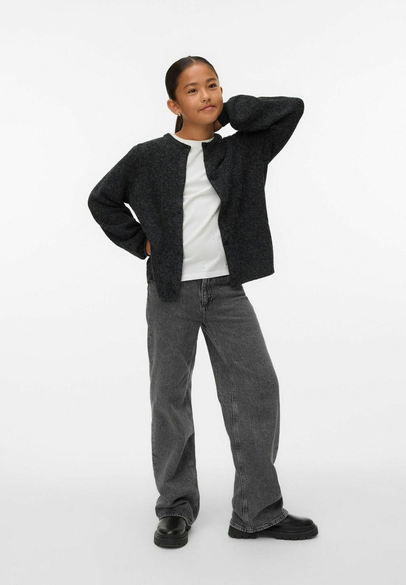 Grauer, langärmeliger, knöpfbarer Cardigan mit lockerem Schnitt, aus einem strukturierten Stoff, kombiniert mit einem weißen T-Shirt und grauen Jeans. Schwarze Schuhe.