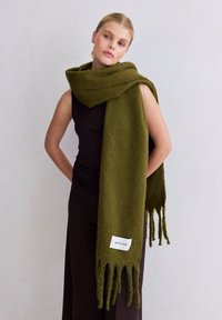 Écharpe en laine verte avec extrémités à franges, portant une étiquette blanche indiquant "ARCTIC FOX". Le modèle la porte drapée sur un haut noir.