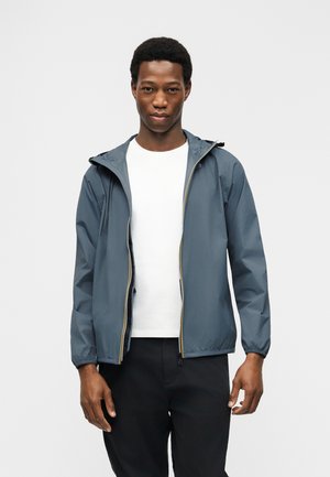 Νεαρός άνδρας φορώντας μπλε μπουφάν με κουκούλα πάνω από λευκό πουκάμισο και μαύρο παντελόνι, στέκεται μπροστά σε απλό ανοιχτό γκρι φόντο.