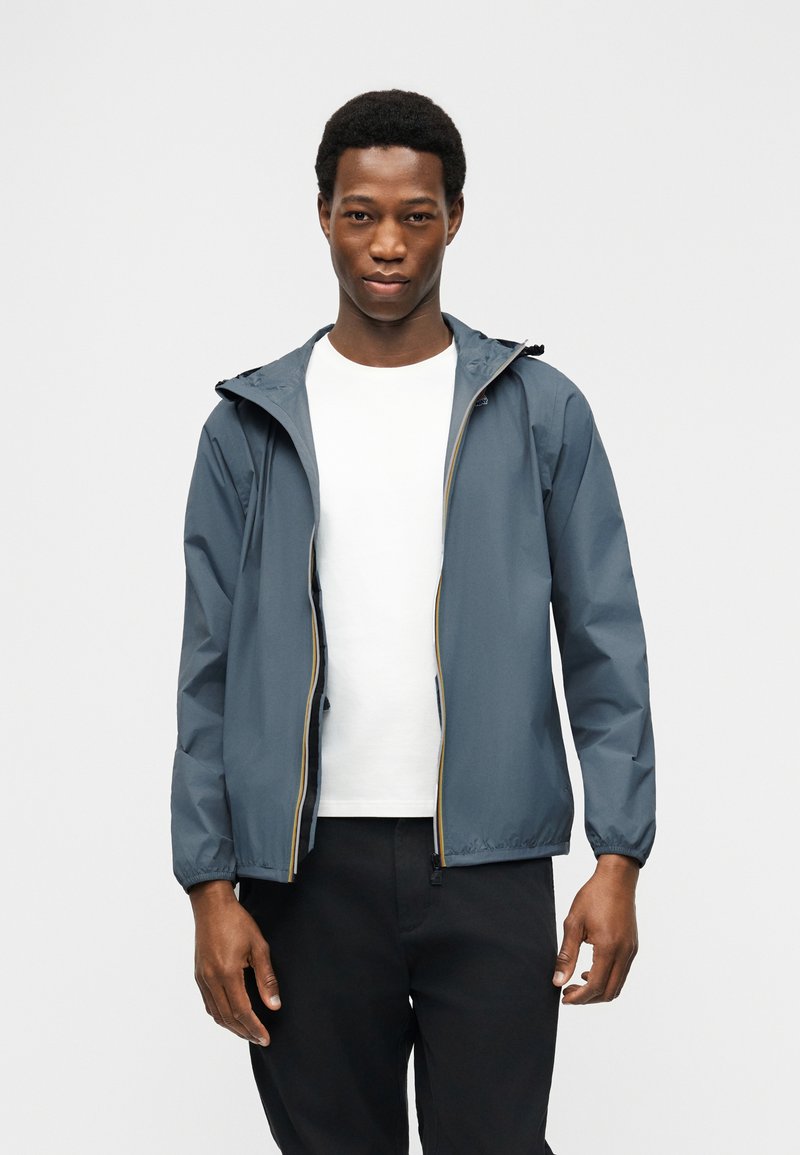 Νεαρός άνδρας φορώντας μπλε μπουφάν με κουκούλα πάνω από λευκό πουκάμισο και μαύρο παντελόνι, στέκεται μπροστά σε απλό ανοιχτό γκρι φόντο.