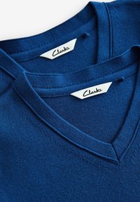 Maglioni v-neck in cotone blu navy impilati insieme, con colletti rigati e etichette "Clarks" in evidenza su tag bianchi.