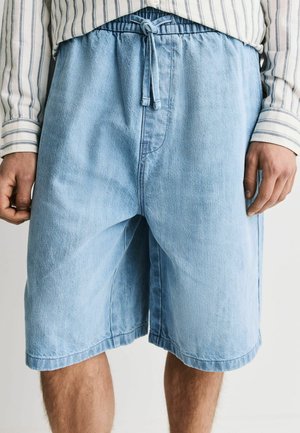 Mann trägt hellblaue Denim-Shorts mit elastischem Bund und Kordelzug, kombiniert mit einem weißen Hemd mit dünnen vertikalen Streifen.