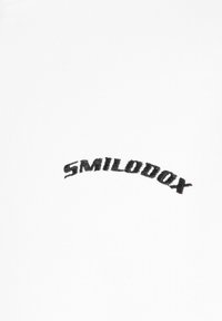 T-shirt bianca con un logo ricamato nero curvo che riporta la scritta "SMILODON" vicino al colletto. Il tessuto ha un aspetto liscio e leggermente strutturato.