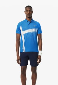 Sininen polo-paita, jossa on valkoinen abstrakti kuvio, valmistettu sileästä kankaasta. Yhdistetty tummansinihin短sareihin ja valkoiseen rannekkeeseen. Lacosten logo näkyvissä.