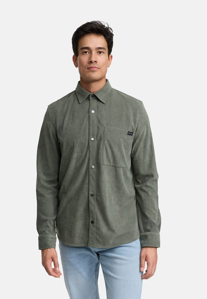 Camisa de pana verde con collar abotonado, bolsillo frontal y botones plateados, con una tela texturizada y mangas largas.