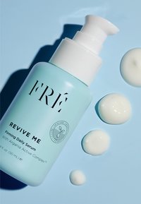 Jasnoniebieska butelka serum "FRÉ Revive Me" z białym dozownikiem, z czarnym napisem i okrągłym logo, obok dwóch małych kropli białego produktu na niebieskim tle.