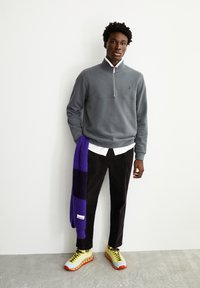 Felpa grigia con zip e colletto, camicia bianca sotto, pantaloni neri e scarpe gialle con accenti rossi. Sciarpa a righe viola e nere.