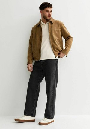 Man draagt een bruine suède jas, wit overhemd met kraag, zwarte jeans met wijde pijpen en witte sneakers, tegen een effen lichte achtergrond.