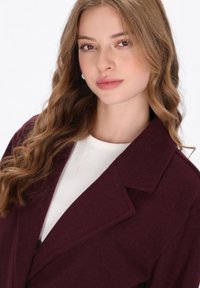 Giovane donna con lunghi capelli castano chiaro ondulati, che indossa orecchini di perle, una maglietta bianca e un cappotto borgogna scuro su uno sfondo bianco.