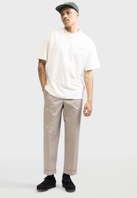 T-shirt en coton beige avec col rond et logo vert. Pantalon gris clair avec ourlets à revers. Chaussures noires avec lacets et surface texturée.