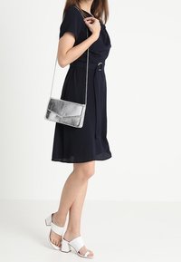 Granatowa sukienka z paskiem w talii, krótkie rękawy; w zestawie z białymi sandałami na słupku i srebrną metaliczną torebką na ramię. Gładkie tekstury.