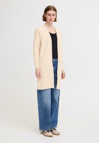 Cardigan long beige à coupe droite et front ouvert, avec deux poches avant. Porté sur un haut noir et un jean bleu à jambes larges.