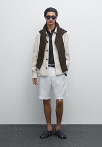 Svetle béžová lněná košile-jacket, černá button-up košile, bílé kraťasy, hnědý pletený svetr přehozený přes ramena, černé sandály, obdélníkové sluneční brýle.
