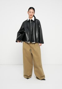 Chaqueta de cuero sintético negra con cuello blanco, de corte oversize y bolsillos frontales. Combinada con pantalones beige anchos y un bolso de mano negro.