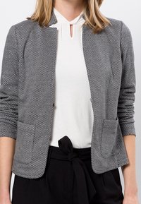 Blazer gris texturé avec un motif tissé, décolleté ouvert, sans col, deux poches avant et manches ajustées. Porté sur un haut blanc.