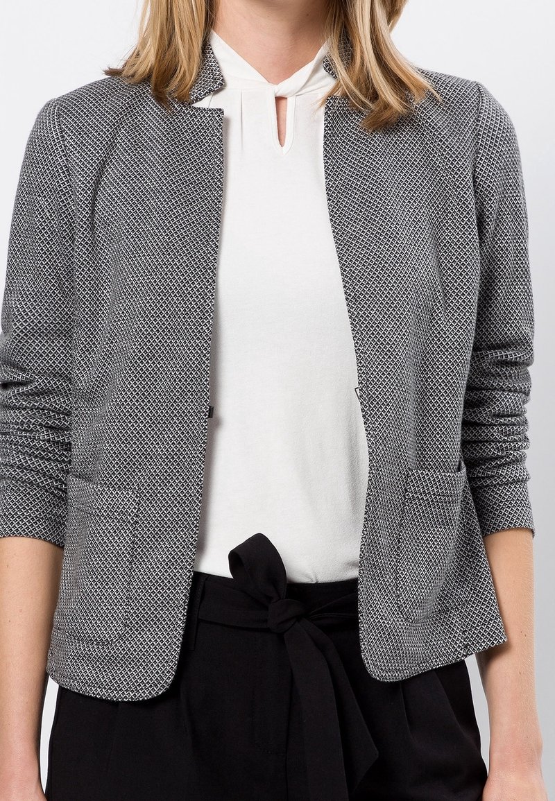 Blazer gris texturé avec un motif tissé, décolleté ouvert, sans col, deux poches avant et manches ajustées. Porté sur un haut blanc.