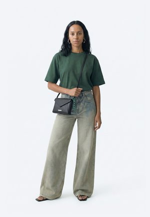 Sieviete ar melniem matiem valkā zaļu T-kreklu, plašas nopelētas džinsas, melnu plecu somu un melnas sandales, stāv pret vienkāršu fona sienu.