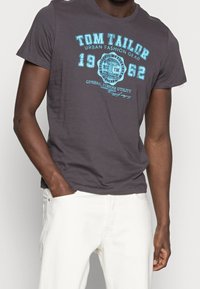 Szary bawełnowy t-shirt z niebieskim napisem "Tom Tailor" i "1962" nadrukowanym na piersi, z okrągłym dekoltem i krótkimi rękawami.